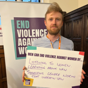 A man holding a placard that says men can end violence against women by listening to women, learning about VAWG, smashing the gender norms that underpin VAWG